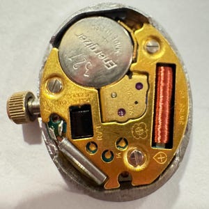 May include: Close-up of a watch movement with a silver case. The internal components include a silver Energizer 321 battery, gold-colored gears, and a red coil. The watch mechanism is designed with small screws and a textured crown.
