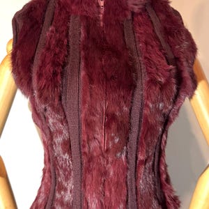 May include: A burgundy sleeveless vest with a high collar and a zipper closure. The vest features vertical panels of knitted fabric and plush fur, creating a textured and layered design. The garment is displayed on a mannequin.
