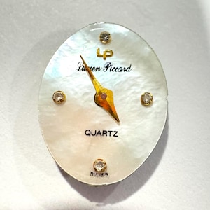 May include: Oval-shaped watch face with a mother-of-pearl background. The watch face features gold hands, four small diamond accents, and the text "Lucien Piccard" and "QUARTZ". The watch face also has the text "SWISS".
