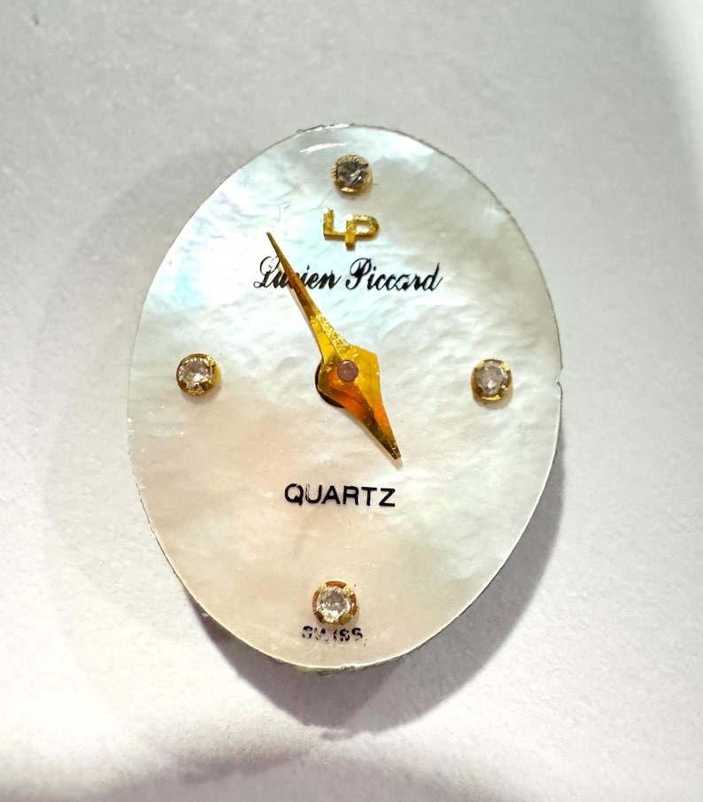 May include: Oval-shaped watch face with a mother-of-pearl background. The watch face features gold hands, four small diamond accents, and the text "Lucien Piccard" and "QUARTZ". The watch face also has the text "SWISS".