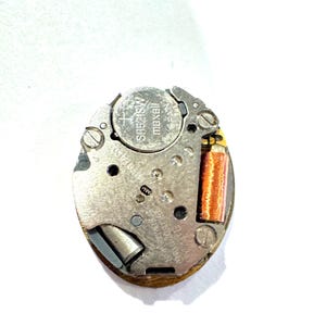 May include: A close-up of a watch movement with a silver metal frame. The watch movement has a Maxell SR521SW battery and a copper-colored coil. The text "NO JEWELS UNADJUSTED" is visible.