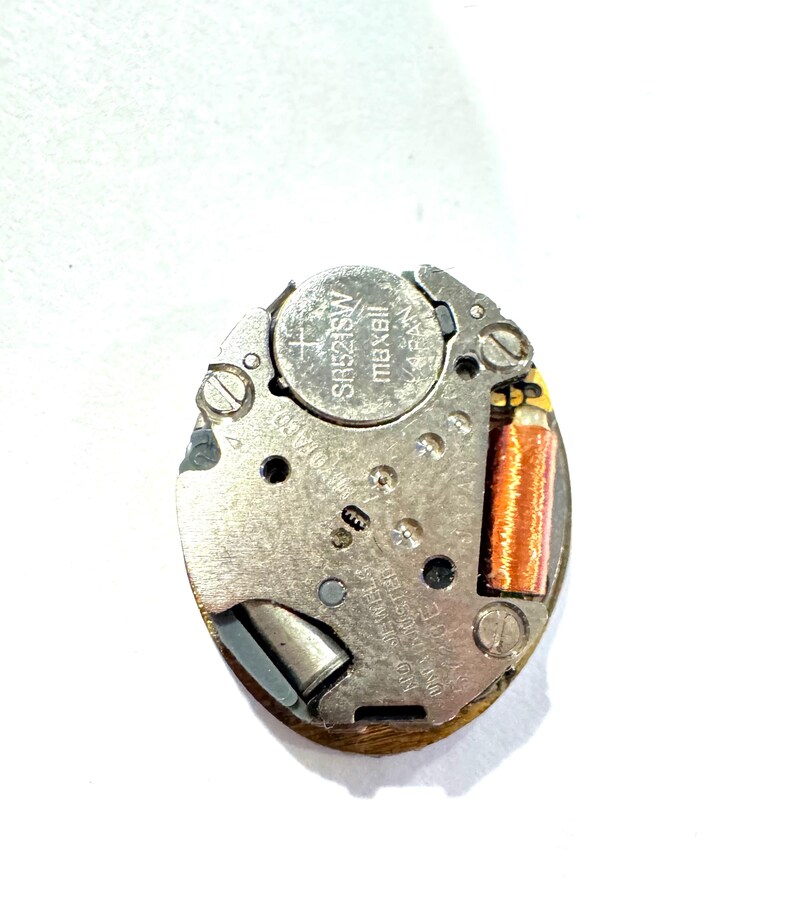 May include: A close-up of a watch movement with a silver metal frame. The watch movement has a Maxell SR521SW battery and a copper-colored coil. The text "NO JEWELS UNADJUSTED" is visible.