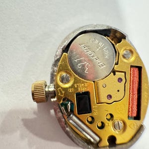 May include: A close-up of a watch movement with a silver-colored metal casing. The movement has a gold-colored base with a silver Energizer 321 battery. The watch movement has a small gold crown and a red coil.