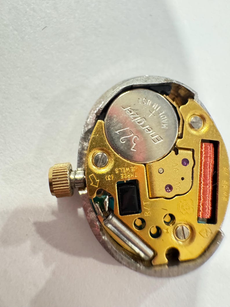 May include: A close-up of a watch movement with a silver-colored metal casing. The movement has a gold-colored base with a silver Energizer 321 battery. The watch movement has a small gold crown and a red coil.
