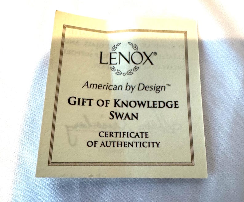 May include: A square, cream-colored Lenox American by Design certificate of authenticity featuring a thin brown border. The text reads Lenox, American by Design, Gift of Knowledge Swan, and Certificate of Authenticity in black lettering.