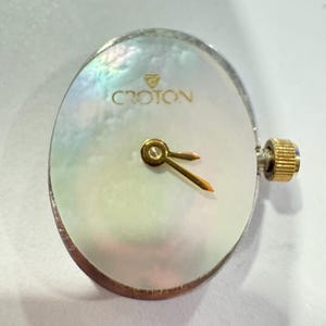 May include: A Croton watch face with a pearlescent, oval-shaped dial. The watch face has gold-colored hands and a gold-colored crown. The brand name "CROTON" is printed in gold lettering at the top of the dial.
