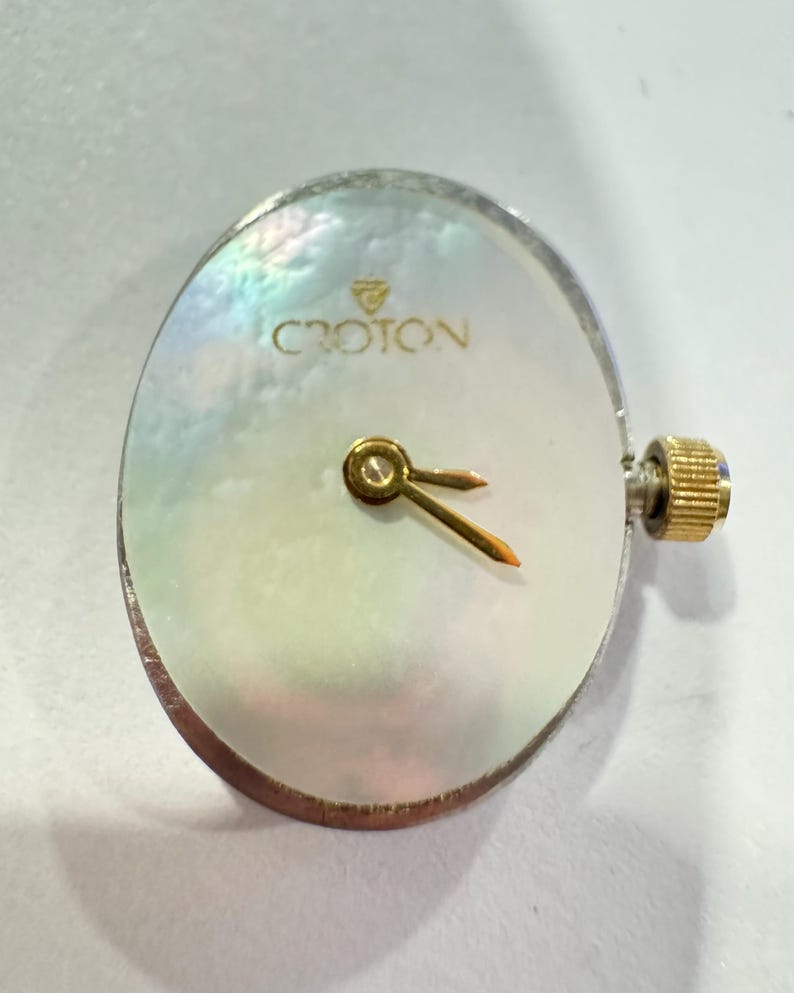 May include: A Croton watch face with a pearlescent, oval-shaped dial. The watch face has gold-colored hands and a gold-colored crown. The brand name "CROTON" is printed in gold lettering at the top of the dial.