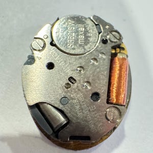 May include: A close-up of a watch movement with a silver-colored metal base. The movement includes a battery labeled "maxell" and a copper-colored coil. The text "UNADJUSTED SWISS" is visible.