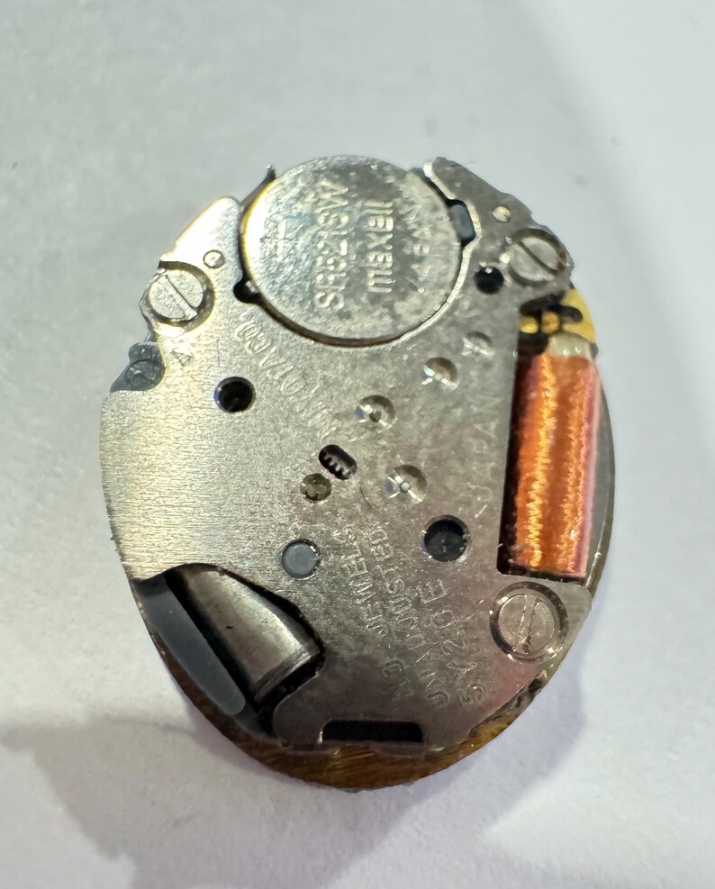 May include: A close-up of a watch movement with a silver-colored metal base. The movement includes a battery labeled "maxell" and a copper-colored coil. The text "UNADJUSTED SWISS" is visible.