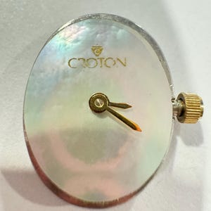 May include: A Croton watch face with a mother-of-pearl dial. The watch face has gold-colored hands and a gold-colored crown. The watch face is oval-shaped and has the brand name "Croton" printed in gold.