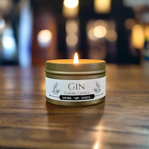 May include: A gold candle tin with a white label that reads "GIN PAIRING CANDLE SWIRL. SIP. SMELL." The candle is lit and the flame is visible.