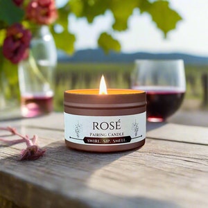 May include: A brown candle with a white label that reads "ROSÉ PAIRING CANDLE SWIRL. SIP. SMELL." The candle is lit and sitting on a wooden table with a glass of red wine in the background.