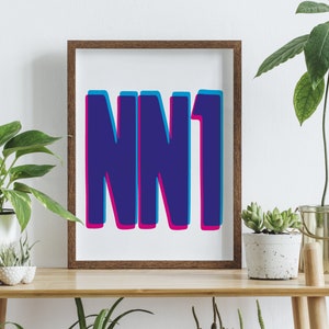 May include: A framed print with the text "NN1" in a bold, blue font with a pink outline. The print is in a brown frame and is sitting on a wooden table.