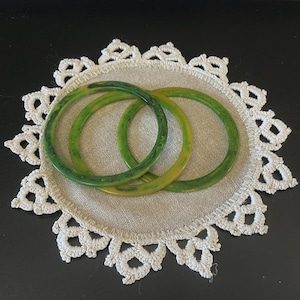 May include: Three circular bangle bracelets in varying shades of green, including dark forest green and light lime green, featuring a smooth, polished finish with subtle marbled patterns throughout the resin material.
