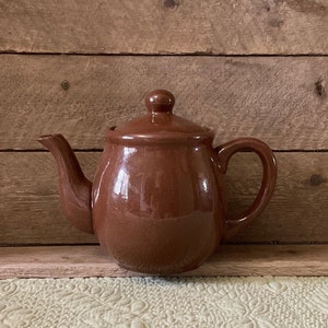 May include: A brown ceramic teapot with a lid and a handle. The teapot is sitting on a wooden surface.