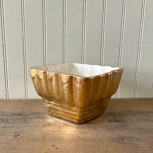 May include: A square-shaped ceramic bowl featuring a metallic gold exterior with a textured, fluted design and a smooth, glossy white interior. The bowl has a wide, open top with scalloped edges and a tapered base, showcasing a reflective finish.
