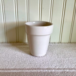 May include: A solid, light beige ceramic plant pot with a smooth, glossy finish. The vessel features a wide, flared rim at the top and a tapered body that narrows toward the base, presenting a clean, simple, and functional design for indoor plants.