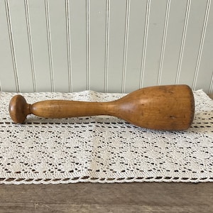 May include: A vintage wooden muddler with a rounded handle and a wide, flat head. The wood has a warm, light brown tone with visible grain and slight imperfections, suggesting age and use. It rests on a cream-colored, crocheted doily.