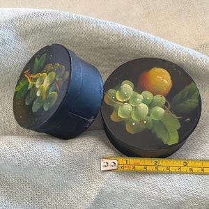May include: Two round black boxes with hand-painted fruit designs featuring green grapes and a yellow peach. A yellow measuring tape is placed next to the boxes, showing a diameter of approximately 3 inches for the circular lids.