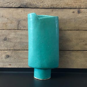 May include: A teal ceramic vase with a unique, asymmetrical design. The vase has a wide opening and a rounded base.