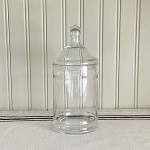 May include: A clear glass apothecary jar with a lid. The jar has a cylindrical body and a rounded base. The lid has a knob on top for easy removal. The jar is sitting on a white surface with a black and white striped fabric.