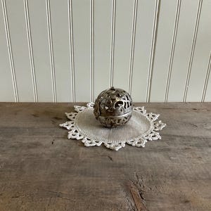 May include: A silver metal sphere with intricate cut-out designs sits atop a cream-colored, crocheted doily. The doily rests on a weathered wooden surface, with a white-paneled wall in the background. The sphere has a small loop at the top.