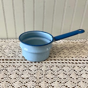 May include: A light blue enamel saucepan with a long, dark blue handle. The rim and handle are a darker shade of blue. The saucepan is sitting on a cream-colored, crocheted doily.
