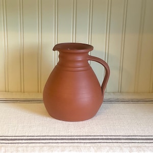 May include: A terracotta-colored pitcher with a rounded body and a handle. The pitcher has a wide opening and a slightly flared rim. The surface is smooth with subtle horizontal lines. The pitcher is a rustic, handmade piece.
