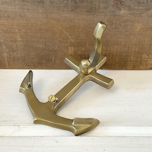 May include: A brass anchor-shaped wall hook. The hook has a classic anchor design with a curved top and a crossbar. The hook is made of brass and has a shiny, metallic finish. The anchor hook is designed to be mounted on a wall.