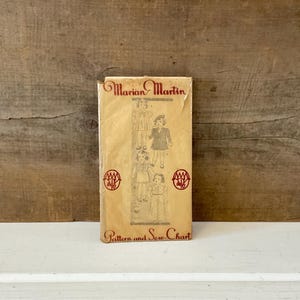 May include: A vintage sewing pattern titled "Marian Martin" with illustrations of children's clothing. The pattern is in a rectangular package with red text and graphics. The background is a wooden surface.