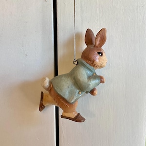 May include: A hanging ornament shaped like a brown rabbit wearing a light blue jacket. The rabbit is depicted in a running pose with dark brown shoes and a white tail. A thin silver string is attached to the top of the jacket for hanging.