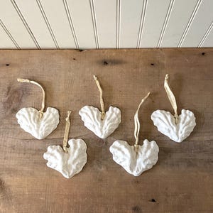 May include: Five white heart-shaped ornaments with textured surfaces and gold-colored hanging ribbons. The ornaments are arranged on a wooden surface, suggesting a rustic or farmhouse style. These decorative hearts are suitable for various occasions.