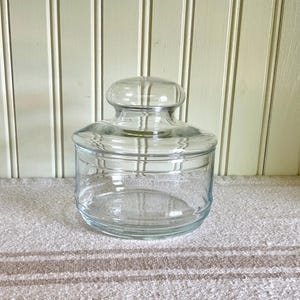 May include: A clear glass storage jar with a wide cylindrical base and a matching glass lid featuring a rounded knob handle. The container has smooth, transparent walls and a thick rim, providing a simple and functional design for storage.