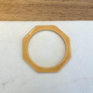 May include: A solid, light tan-colored bangle bracelet with a distinct octagonal outer shape and a circular inner opening. The surface of the bangle features a smooth, matte finish with subtle, natural-looking variations in the light brown color tone.