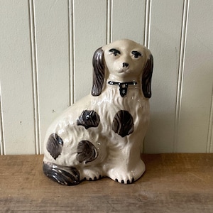 May include: A ceramic dog figurine in a seated position with a glossy cream-colored finish. The dog features dark brown painted ears, tail, and scattered spots across its body. It wears a simple black collar around its neck with painted details.