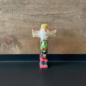 May include: A small, colorful totem pole figurine. The top features a yellow-headed owl with white feathers and red accents. Below, the pole displays green, black, and red carved figures. The figurine is set against a wooden background.