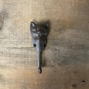 May include: A cast iron cat-shaped wall hook in a dark brown finish. The hook features a detailed cat face at the top with a single hook extending downwards. The hook is mounted on a wooden surface.