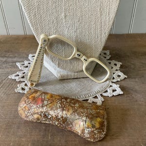 May include: A pair of off-white folding lorgnette glasses featuring a handle decorated with a row of small silver-toned studs. The glasses are accompanied by a rectangular, translucent case with a gold-toned metallic wire mesh overlay.