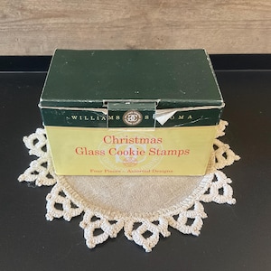 May include: A rectangular box for Williams Sonoma Christmas Glass Cookie Stamps. The box features a dark green lid and a light yellow base with red text that reads Christmas Glass Cookie Stamps and Four Pieces - Assorted Designs.