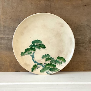 May include: A round, off-white ceramic plate with a hand-painted design of a green pine tree. The tree's branches and needles are detailed against the plate's neutral background. The plate is a decorative item.