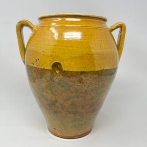 May include: A large, antique ceramic jar with two handles. The upper portion is a glossy yellow, while the lower half is a mottled mix of brown, green, and red. The jar has a wide mouth and a rounded body.