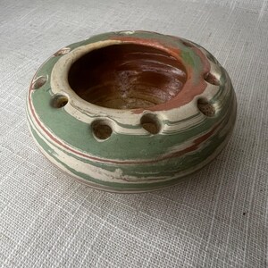 May include: A round, ceramic vase with a wide opening and several small holes around the rim. The vase features a swirl pattern of green, white, and reddish-brown colors. The interior is a darker brown.
