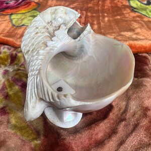 May include: A white seashell with a textured surface, shaped like a fish with an open mouth. The shell is mounted on a white base.