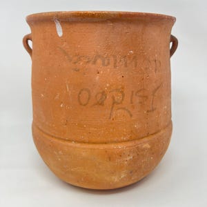 May include: A terracotta-colored ceramic pot with two small handles and a rounded base. The pot has a slightly rough texture and appears to be handmade. The pot has some writing on the side. The pot is likely used for storage or decoration.