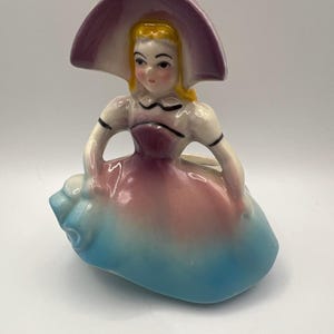 May include: A ceramic figurine of a person wearing a wide-brimmed purple hat, a white bodice with black trim, and a long, flared skirt that transitions from a pink midsection to a bright blue base, featuring yellow hair and painted facial features.