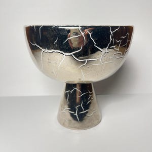 May include: A silver-toned pedestal bowl featuring a reflective, metallic finish overlaid with a distinct white crackle pattern. The bowl has a wide, rounded upper basin that tapers down into a sturdy, conical base, creating a chalice-like shape.