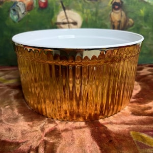 May include: A gold-colored ceramic baking dish with a white interior and a fluted design. The dish is round and has a wide rim.