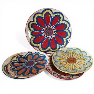 May include: A set of decorative ceramic plates featuring a large floral design. The plates have a white background with a red, blue, yellow, and orange flower pattern. The plates are round and oval-shaped, with a red border.