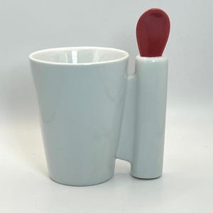 May include: A white ceramic mug with a smooth, glossy finish and a wide, open top. Attached to the side of the mug is a vertical, cylindrical handle that features a bright red, spoon-shaped top element integrated into the design of the vessel.