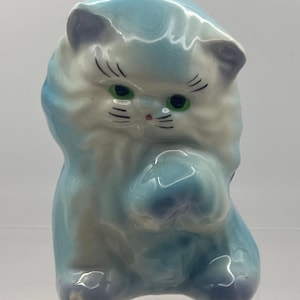 May include: A glossy ceramic cat figurine featuring a light blue and white marbled glaze. The cat has painted green eyes, a small pink nose, and black whiskers. It is depicted in a seated position with one paw raised near its face.